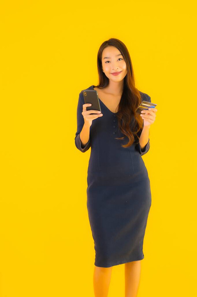 Portrait beautiful young asian woman with smart mobile phone or cell phone and credit card yellow isolated background - Fotó, kép