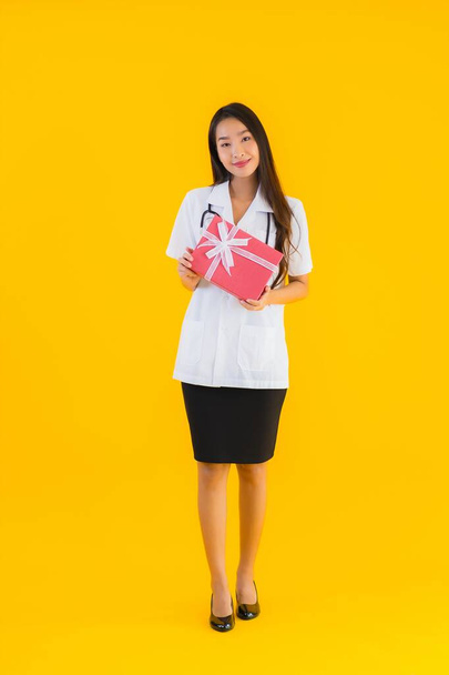 Portrait beautiful young asian doctor woman show red gift box on yellow isolated background - Photo, Image