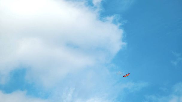 凧は雲の中を空を飛んでいる。コンセプト。白い雲と高くそびえる虹色の凧、自由と子供時代の概念を持つ夏の青い空の下の景色. - 写真・画像