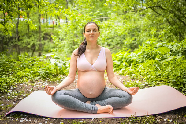 Piękna młoda ciężarna kobieta ćwiczy jogę w parku na świeżym powietrzu. Siedząc i relaksując się na różowej macie do jogi. Aktywny przyszły styl życia matki sportu. Koncepcja zdrowej ciąży - Zdjęcie, obraz