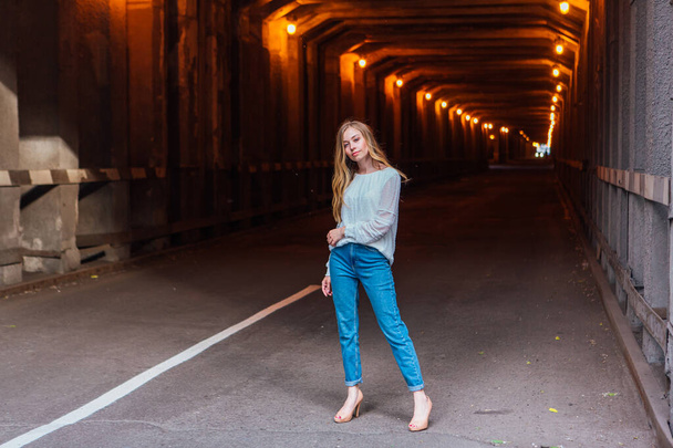 Young woman with long blond hair standing in a dark industrial tunnel with lights - Foto, Imagen