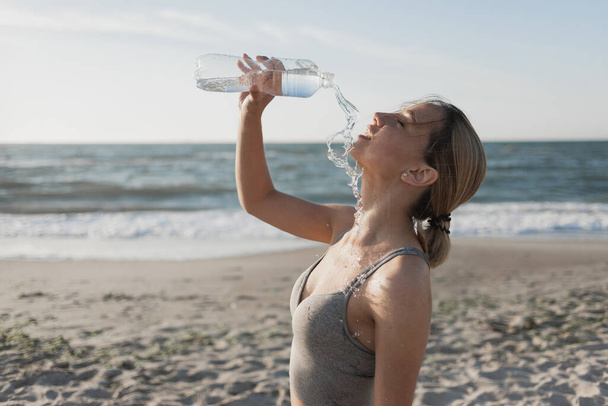 mooie jonge vrouw drinkt water na een training. Moe giet vloeistof over zichzelf. Lekker. Drijf je dorst weg. Oceaan. Plaats voor tekst - Foto, afbeelding