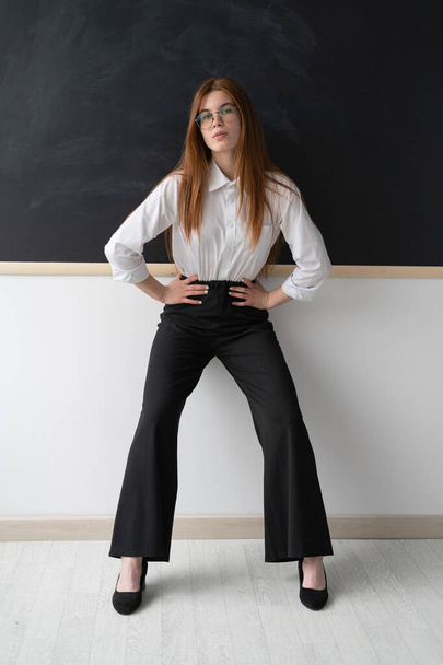 Beautiful young high school teacher in the office. She is standing in front of a clean black board. She is wearing a white womens shirt and black trousers, as well as black pointed toe shoes. Her - Photo, Image