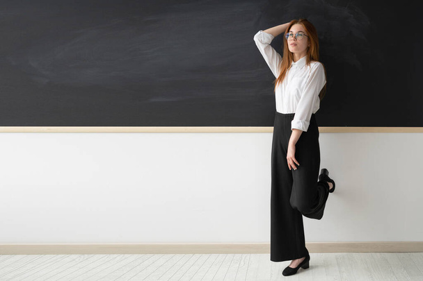 学校では非常に美しい若い女性教師。彼女はオフィスの黒い黒板の前に立っている。女の子は白いシャツと黒いパンツを着ている。彼女は若いヒールの高い靴を持っています - 写真・画像