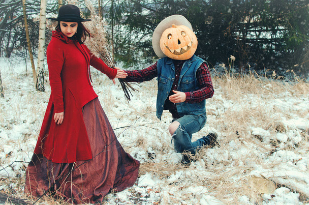 Espantalho de Halloween com uma abóbora na cabeça dá a uma bruxa um buquê de ervas secas em pé em um joelho em uma clareira da floresta
 - Foto, Imagem
