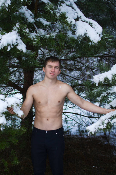 jongeman zonder shirt in het winterwoud staat onder een boom - Foto, afbeelding