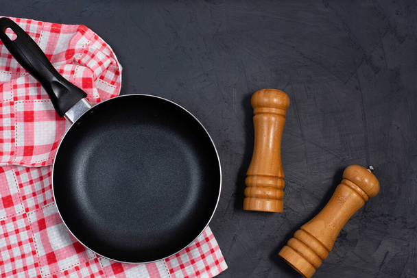 Κενό μαύρο τηγάνι ή τηγάνι με πιπέρι και θαλασσινό αλάτι σε μια κόκκινη χαρτοπετσέτα καρό σε μαύρο τραπέζι. Στο πάνω μέρος. Αντιγραφή χώρου - Φωτογραφία, εικόνα