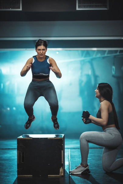 young fit woman is doing hard exercise at the gym. She is jumping on the boxes supporting by female coach. - Foto, imagen