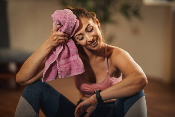 Νεαρή γυναίκα ξεκουράζεται κατά τη διάρκεια της αθλητικής εκπαίδευσης στο σαλόνι στο σπίτι. Απολαμβάνει την πρωινή λιακάδα, σκουπίζοντας το μέτωπό του με μια πετσέτα. - Φωτογραφία, εικόνα