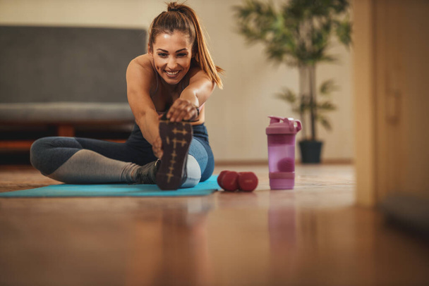 Mujer sonriente joven está haciendo ejercicios de estiramiento en la sala de estar en la alfombra del piso en casa bajo el sol de la mañana. - Foto, Imagen