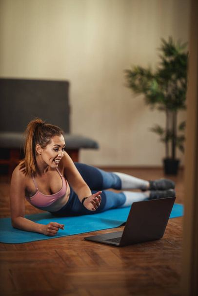 Młoda uśmiechnięta kobieta nagrywa poradnik internetowy i ćwiczy sport w salonie na macie przed laptopem. - Zdjęcie, obraz