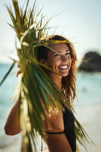 ヤシの葉の下の熱帯のビーチでポーズをとって楽しんでいる魅力的な若い女性の肖像画. - 写真・画像
