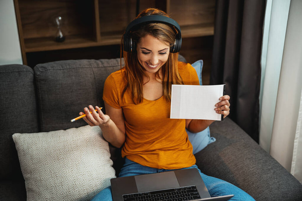 Söpö nuori naispuolinen opiskelija käyttää kannettavaa tietokonetta ja kuulokkeita ja oppimista kotona. - Valokuva, kuva