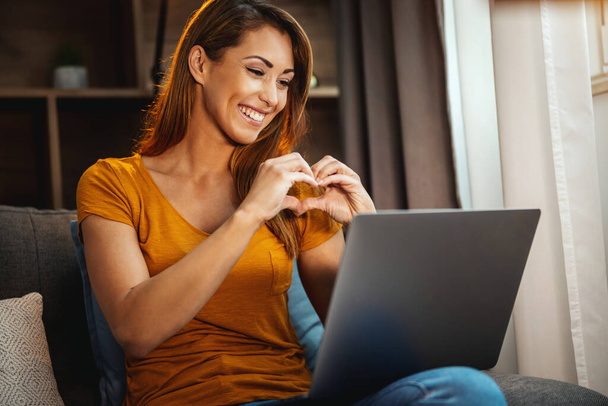 Atrakcyjna młoda kobieta siedząca na kanapie ze skrzyżowanymi nogami i korzystająca z laptopa, aby nagrać wideo czat z kimś w domu. - Zdjęcie, obraz