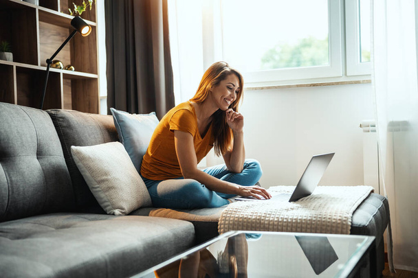 Attraktive junge Frau sitzt auf dem Sofa und benutzt ihren Laptop zu Hause. - Foto, Bild