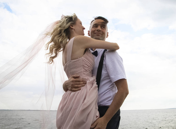 Creative hipster wedding by the sea. The bride is in a pink dress and veil, the groom is in beautiful trousers with suspenders and a youth shirt. Madness, fun and an unusual approach to marriage - Photo, Image