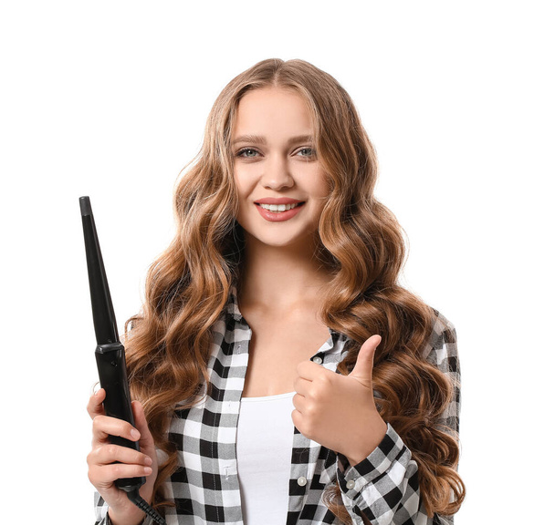 Beautiful young woman with curling iron for hair showing thumb-up against white background - Photo, Image