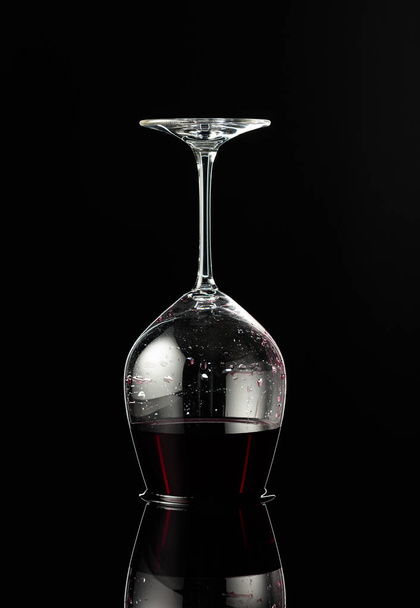 Ανεστραμμένο ποτήρι κρασιού με κόκκινο κρασί σε μαύρο ανακλαστικό φόντο. Έννοια της οινοποίησης. - Φωτογραφία, εικόνα