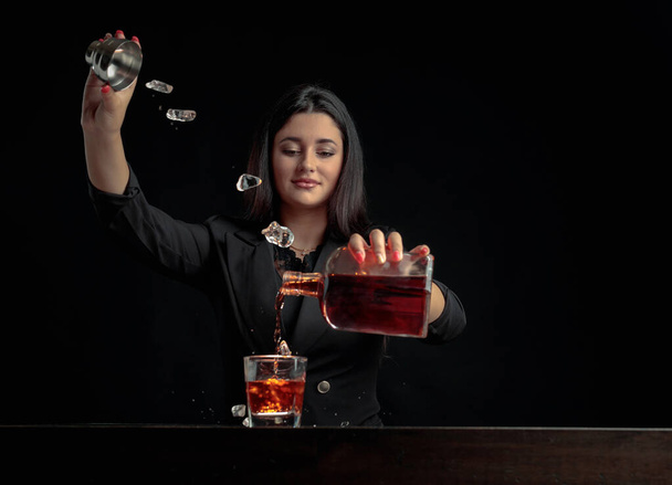 Приваблива дівчина-бармен п'є алкогольний напій у склянку і кидає шматочки льоду. Красива брюнетка в чорному готує коктейль. Копіювати простір
. - Фото, зображення