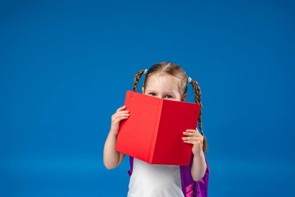 charming mischievous little girl with glasses smiles at the camera and hides behind a book, having fun during school classes on a blue background. Back to school. - Zdjęcie, obraz
