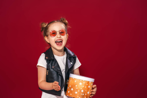 Stylish charming little girl in fashionable clothes with a naughty hairstyle in sunglasses with a gift box shows the V sign A child is celebrating a holiday and having fun, posing on a red background. - Photo, image