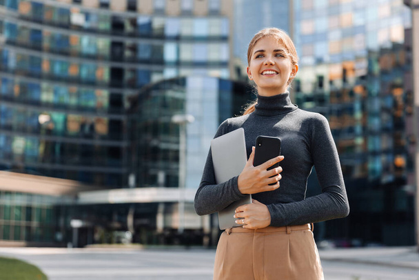Porträt einer jungen blonden Geschäftsfrau mit einem Lächeln, die einen Laptop und ein Mobiltelefon in der Hand hält, steht auf der Straße, im Geschäftsviertel der Stadt, vor dem Hintergrund eines Geschäftszentrums - Foto, Bild