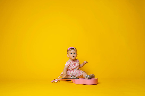 Cute little baby 1 year old, in a pink dress with a floral print and a headband, playing guitar on a yellow background. Copy space. Previously, the development of the child. The teaching of music. - Zdjęcie, obraz