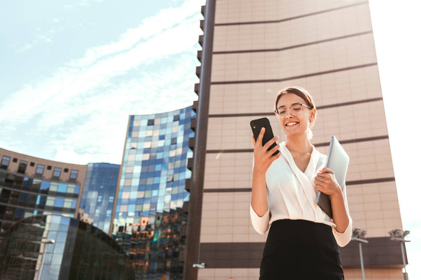 Smiling business woman in glasses and a blouse holds a laptop and uses the phone outdoors in a modern city against the background of a business center, on a sunny day - Foto, afbeelding