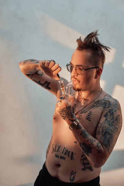 Hombre sin camisa con cuerpo tatuado y corte de pelo raro encendiendo cannabis en la pipa - Foto, imagen