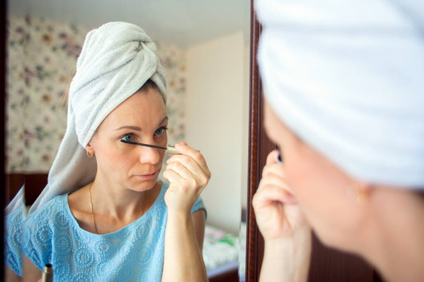 Una mujer con la cabeza lavada en una toalla maquilla su habitación por la mañana. Chica pinta pestañas con rímel
 - Foto, Imagen