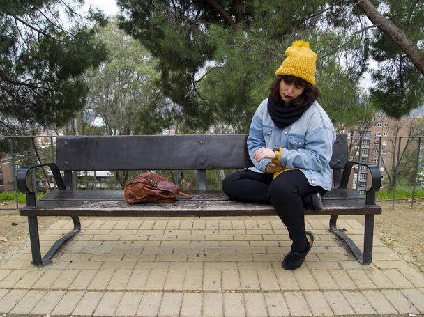 Mladá žena se žlutou vlněnou čapkou sedící na lavičce a dívající se na hodinky čekající na někoho. Podzimní městský koncept. - Fotografie, Obrázek