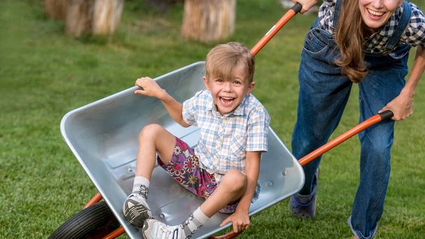 Happy little boy having fun in a wheelbarrow in domestic garden on warm sunny day. Active outdoors games for kids in summer. - Photo, Image