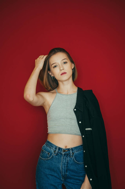 Attractive girl in jeans and t-shirt stands on a red background and poses at the camera with a serious face, straightens her hair, isolated. Serious model in casual clothes isolated on red background - Zdjęcie, obraz