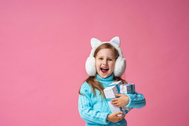 Merry Christmas! happy little girl in fur headphones with gifts in her hands on a pink background. The child is happy and enjoys the holiday. Christmas gift. Advertizing. Christmas eve and the spirit - Zdjęcie, obraz