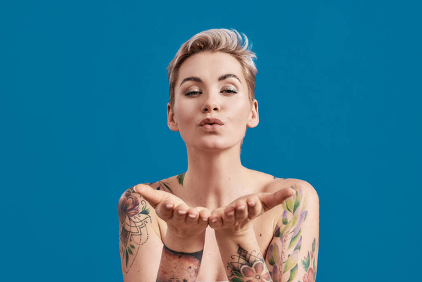 Portrait of a young attractive half naked tattooed woman with perfect skin blowing kiss to camera isolated over blue background - Foto, afbeelding