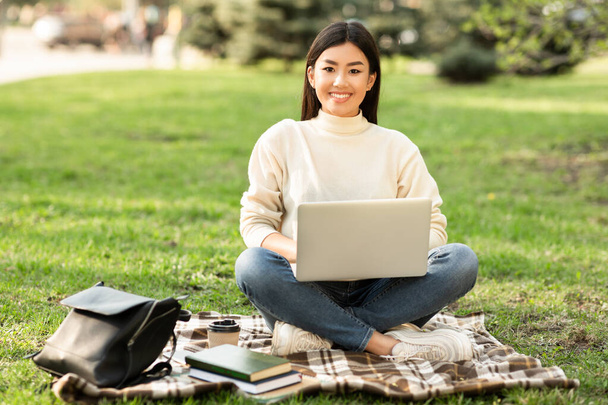 Aasian nainen käyttää kannettavaa tietokonetta, istuu puistossa, katselee kameraa - Valokuva, kuva