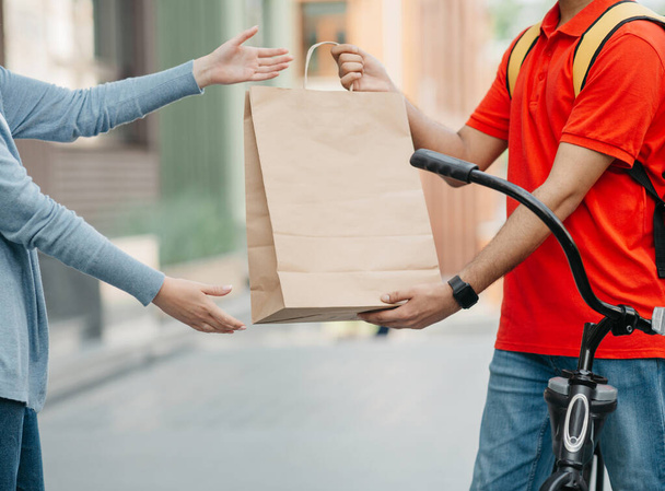 Online shopping and delivery by courier on bike. Man with backpack gives paper bag to client - Photo, Image