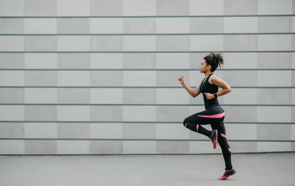 Corredor en pista después del entrenamiento. Chica afroamericana en ropa deportiva y con rastreador de fitness, trotando - Foto, Imagen