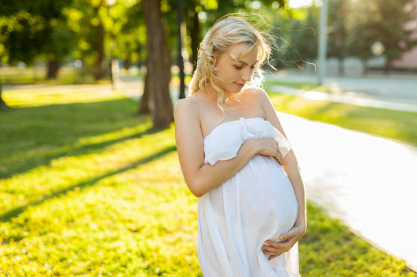 Ritratto di donna incinta felice e orgogliosa in abito bianco che si guarda la pancia, in un parco nella soleggiata giornata estiva con una calda luce posteriore sullo sfondo - Foto, immagini