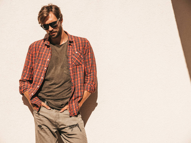 Portrait of handsome stylish hipster lambersexual model.Man dressed in checkered shirt. Fashion male posing in the street near white wall outdoors in sunglasses - Photo, image
