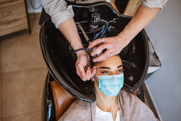 A young woman in a medical mask washes her hair in a barbershop during the covid-19 coronavirus pandemic. Reopening with security measures of Hairdressers in the Covid-19 pandemic - Foto, Imagem