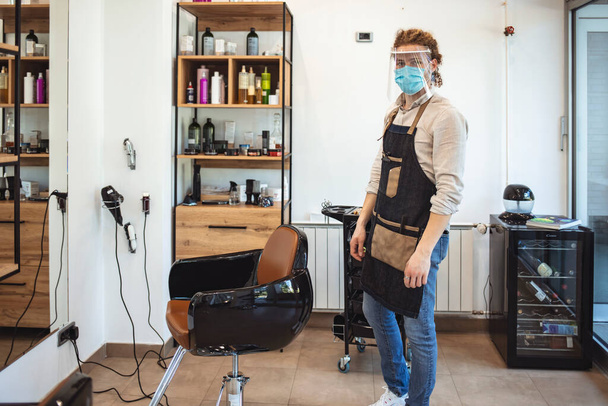 Hair stylist portrait in working place during pandemic isolation, he wears protective equipment. Man standing in his salon. Professional hairdresser with all the hairdressing accessories tied to her waist. - Foto, imagen