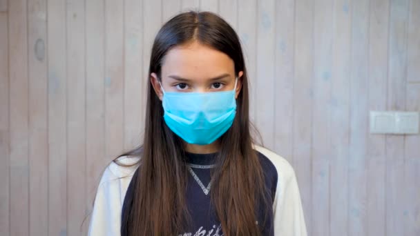 Teenage dívka na sobě lékařské ochranné masky při pohledu do kamery. Covid-19, epidemie koronaviru. Portrét krásné brunetky teen dívka v ochranné masky přes světle šedé pozadí stěny. - Záběry, video