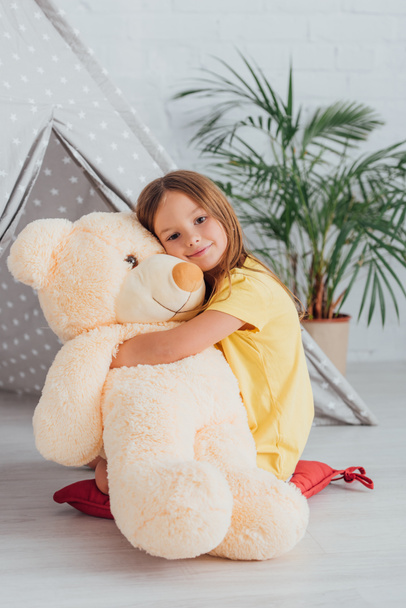 girl in white t-shirt hugging teddy bear while sitting on floor near play tent - Photo, Image