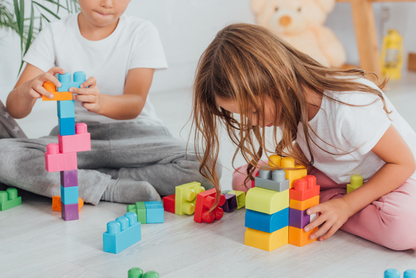 bijgesneden uitzicht van jongen spelen met bouwstenen in de buurt van zus terwijl zitten op de vloer in pyjama 's - Foto, afbeelding