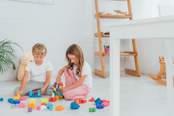 selective focus of focused brother and sister in pajamas sitting on floor and playing with building blocks - Foto, Bild
