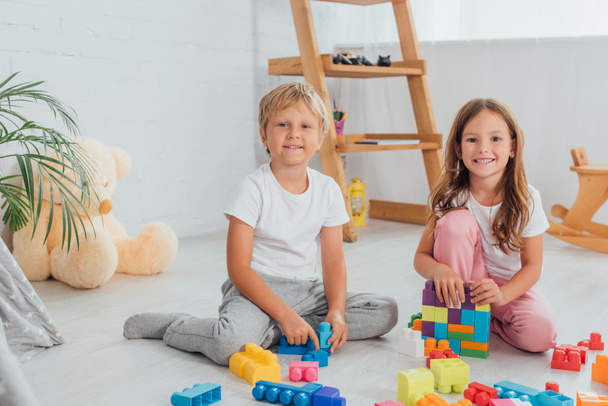 excited brother and sister in pajamas sitting on floor near building blocks and looking at camera - 写真・画像