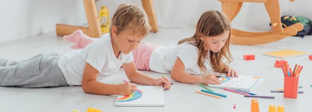 irmão concentrado e irmã de pijama deitado no chão e desenho com canetas de feltro em cadernos
 - Foto, Imagem