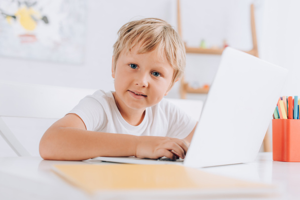 selektywna ostrość dziecka w białej koszulce patrzącego w kamerę siedzącego przy stole i korzystającego z laptopa - Zdjęcie, obraz