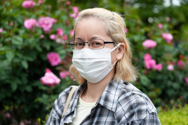 Gemengde ras blonde vrouw van middelbare leeftijd met een bril met wit chirurgisch masker. Bescherming tegen coronavirus (COVID-19) en andere infectieziekten. Buiten. Kijkend naar de camera. - Foto, afbeelding